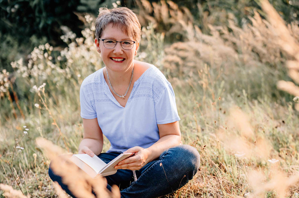 Marion Schmitz sitzt umgeben von hohen Gräsern auf der Wiese mit einem Buch in der Hand